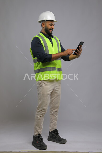 Using engineering applications for architectural projects, integrating work with technique and technology, portrait of a smiling Saudi Arabian Gulf architect wearing a helmet and protective vest, using a mobile phone, urban growth and development in Saudi Arabia, full-length body portrait, gray background