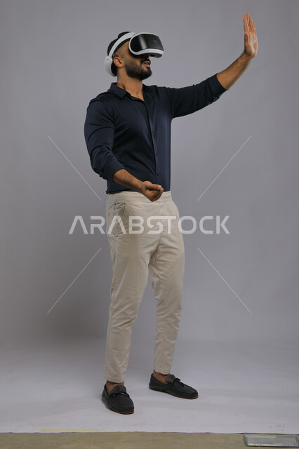The concept of developing 3D games using modern electronic technologies, an attempt to touch and move an element, a portrait of a smiling Saudi Arabian Gulf man wearing casual clothes and smart VR glasses, interaction and integration with virtual reality, full-length body image, gray background