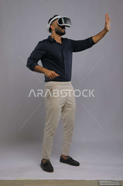 استخدام حاسة اللمس لتحريك عنصر ما, مشاهدة الأفلام ومحاكاة العالم الخيالي, بورتريه لشاب عربي سعودي خليجي مبتسم يرتدي الزي الكاجوال ونظارة في آر ثلاثية الأبعاد, التفاعل والاندماج مع ألعاب الواقع الافتراضي,  صورة الجسم بالطول الكامل, خلفية رمادية.