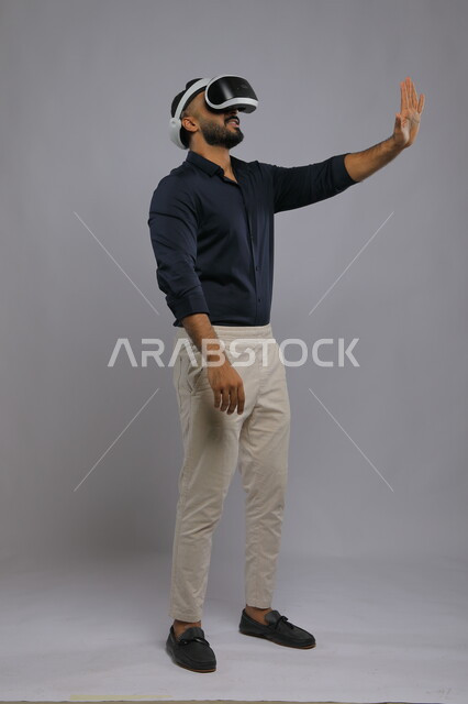 Interacting and merging with virtual reality games, simulating the imaginary world and trying to touch an item, a portrait of a smiling Saudi Arabian Gulf man wearing casual clothes and smart 3D VR glasses, advanced technologies and devices, full-length body image, gray background