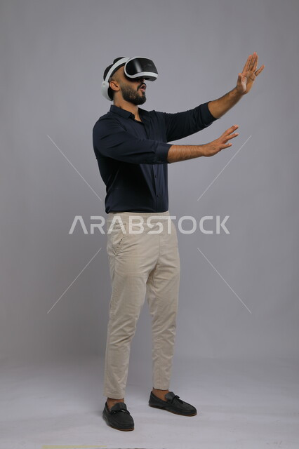 Simulating the imaginary world and trying to touch an item, gestures of amazement and enjoyment, portrait of a Saudi Gulf Arab man wearing casual clothes and 3D VR glasses, interaction and integration with virtual reality games, advanced technologies and devices, full-length body image, gray background