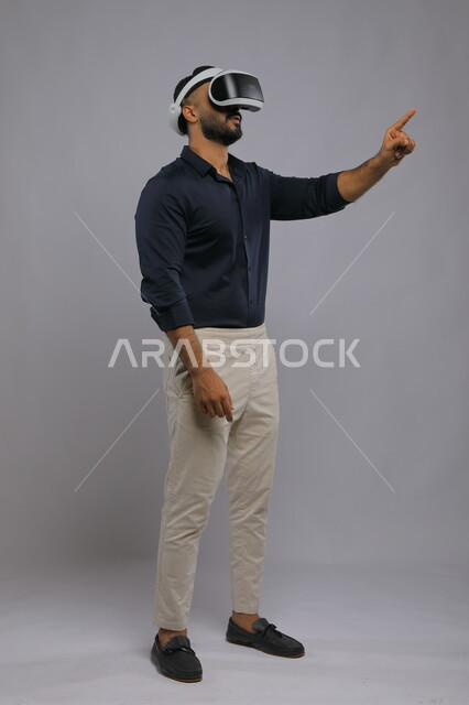 محاولة النقر والضغط على عنصر ما,مفهوم تطوير الألعاب ثلاثية الأبعاد بتقنيات الكترونية حديثة, بورتريه لرجل عربي سعودي خليجي مبتسم يرتدي الزي الكاجوال ونظارة في آر الذكية, التفاعل والاندماج مع  الواقع الافتراضي, صورة الجسم بالطول الكامل, خلفية رمادية.