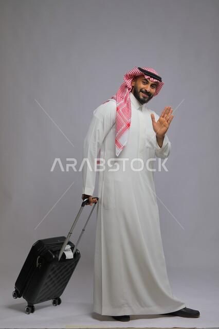قضاء وقت ممتع في السياحه بالعطلة الصيفيه ،النظر إلى الكاميرا بإيماءات الوداع للذهاب، الاستعداد للانطلاق و بدء الرحلة، بورتريه لشاب عربي خليجي سعودي مسرور يرتدي الشماغ والثوب التقليدي يمسك حقيبة السفر ،صورة الجسم بالطول الكامل ،خلفية رمادية