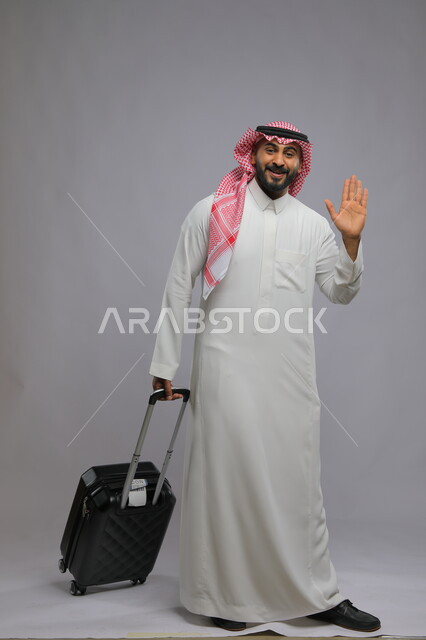 Looking at the camera with farewell gestures to go, preparing to set off and start the trip, spending a good time in tourism during the summer vacation, portrait of a happy Saudi Gulf Arab young man wearing a shemagh and traditional thobe, holding a travel bag, full-length body portrait, gray background