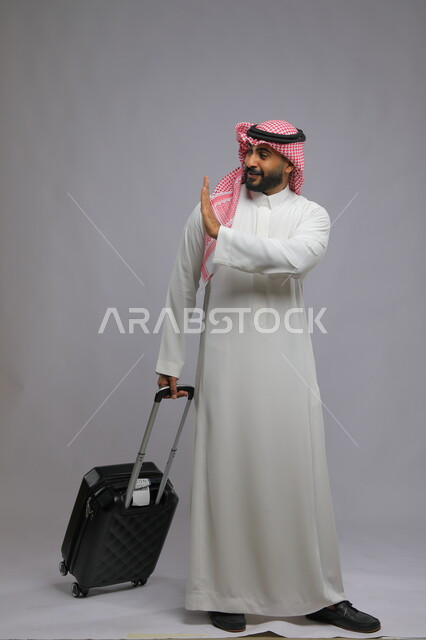 ايماءات الوداع للذهاب، الاستعداد للانطلاق و بدء الرحلة، قضاء وقت ممتع في السياحه بالعطلة الصيفيه، الوصول على موعد الطائرة ،بورتريه لشاب عربي خليجي سعودي يرتدي الشماغ والثوب التقليدي يمسك حقيبة السفر ،صورة الجسم بالطول الكامل ، خلفية رمادية