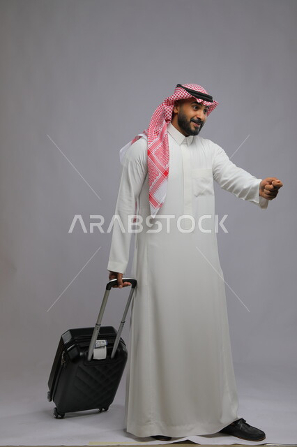 تقديم جواز السفر لموظف الريسبشين للتأكد من فعاليته، قضاء وقت ممتع في السياحه بالعطلة الصيفيه، بورتريه لشاب عربي خليجي سعودي مبتسم يرتدي الشماغ والثوب التقليدي يمسك بيده حقيبة سفر ،صورة الجسم بالطول الكامل ، خلفية رمادية