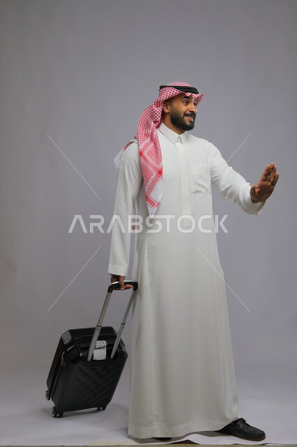 رفع اليد بايماءات الابتعاد و طلب التوقف، عدم الحاجة للمساعدة، قضاء وقت ممتع في السياحه بالعطلة الصيفيه، بورتريه لشاب عربي خليجي سعودي مبتسم يرتدي الشماغ والثوب التقليدي يمسك بيده حقيبة سفر ،صورة الجسم بالطول الكامل ، خلفية رمادية