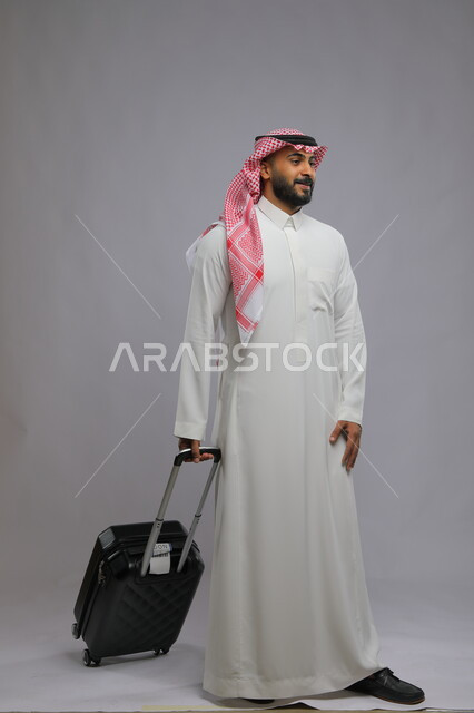 الانتظار للكشف والتفتيش على الحقائب ، قضاء وقت ممتع في السياحه بالعطلة الصيفيه، عدم التأخر على موعد الطائرة ،بورتريه لشاب عربي خليجي سعودي يرتدي الشماغ والثوب التقليدي يمسك بيده حقيبة السفر ،صورة الجسم بالطول الكامل ، خلفية رمادية