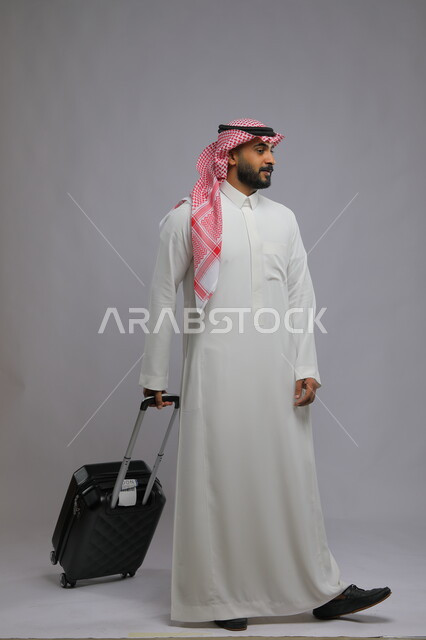 الاستعداد للانطلاق و بدء الرحلة، قضاء وقت ممتع في السياحه بالعطلة الصيفيه، الوصول على موعد الطائرة ،بورتريه لشاب عربي خليجي سعودي يرتدي الشماغ والثوب التقليدي يقوم بجر حقيبة السفر خلفه ،صورة الجسم بالطول الكامل ، خلفية رمادية