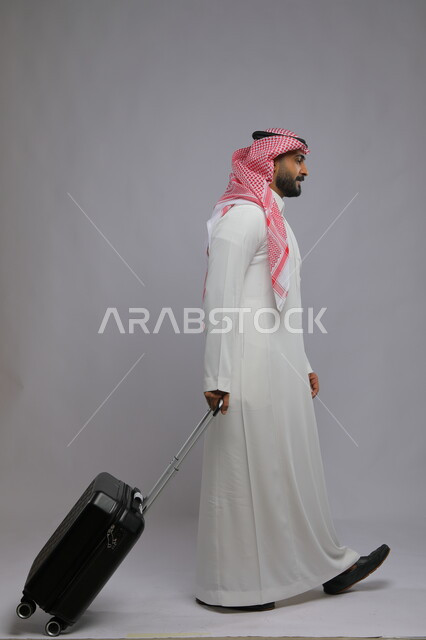 الوصول على موعد الطائرة ،الذهاب والمشي إلى المطار، قضاء وقت ممتع في السياحه بالعطلة الصيفيه، بورتريه لشاب عربي خليجي سعودي يرتدي الشماغ والثوب التقليدي يقوم بجر حقيبة السفر خلفه ،صورة الجسم بالطول الكامل ، خلفية رمادية