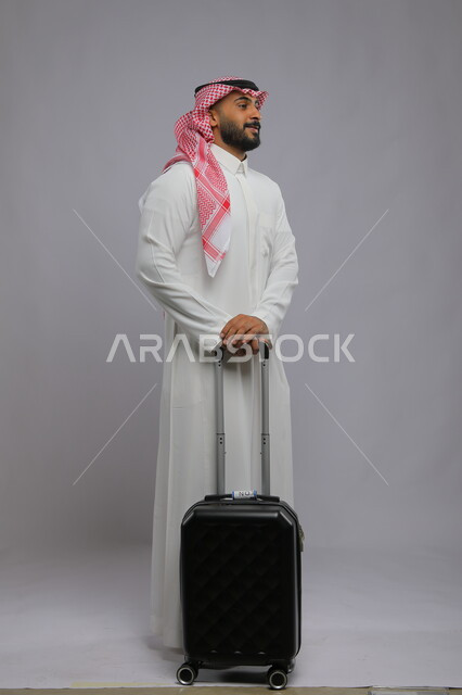 الوقوف باستقامة و انتظار موعد الطائرة ، قضاء وقت ممتع في السياحه بالعطلة الصيفيه، النظر إلى  شيء ما، بورتريه لشاب عربي خليجي سعودي مبتسم يرتدي الشماغ والثوب التقليدي يمسك بيده حقيبة سفر ،صورة الجسم بالطول الكامل ، خلفية رمادية