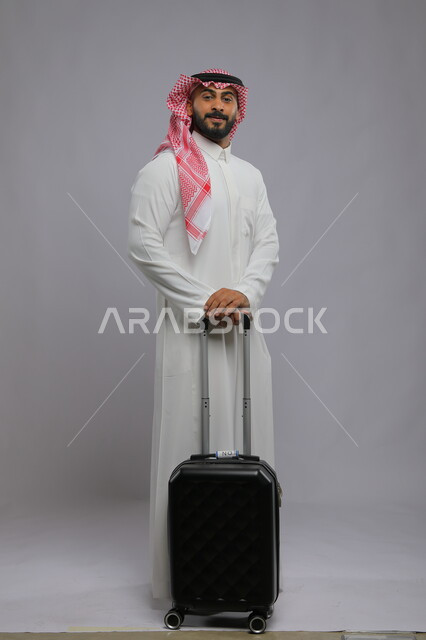 الوقوف باستقامة والنظر إلى الكاميرا ،انتظار موعد الطائرة ، قضاء وقت ممتع في السياحه بالعطلة الصيفيه، بورتريه لشاب عربي خليجي سعودي مبتسم يرتدي الشماغ والثوب التقليدي يمسك بيده حقيبة سفر ،صورة الجسم بالطول الكامل ، خلفية رمادية