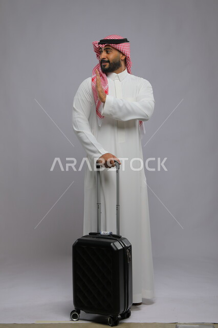 الاستمتاع في السياحه بأوقات الفراغ، رفع اليد بايماءات الشكر وعدم الحاجه للمساعده، بورتريه لشاب عربي خليجي سعودي مبتسم يرتدي الشماغ والثوب التقليدي يمسك بيده حقيبة سفر ،صورة الجسم بالطول الكامل ، خلفية رمادية