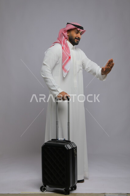 رفع اليد بايماءات طلب التوقف والابتعاد، الاستمتاع  في السياحه بأوقات الفراغ، بورتريه لشاب عربي خليجي سعودي مبتسم يرتدي الشماغ والثوب التقليدي يمسك بيده حقيبة سفر ،صورة الجسم بالطول الكامل ، خلفية رمادية