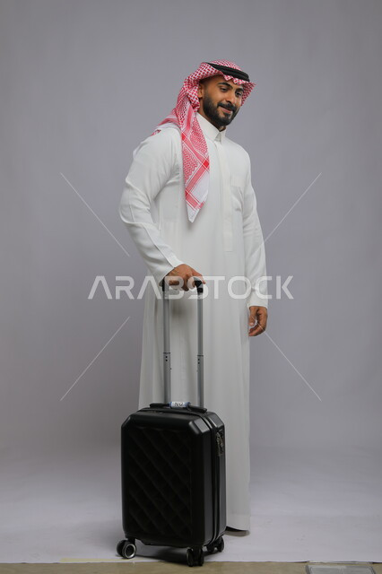 الوقوف بإيماءات السرور وانتظار موعد الطائرة ، قضاء وقت ممتع في السياحه بالعطلة الصيفيه، بورتريه لشاب عربي خليجي سعودي مبتسم يرتدي الشماغ والثوب التقليدي يمسك بيده حقيبة سفر ،صورة الجسم بالطول الكامل ، خلفية رمادية