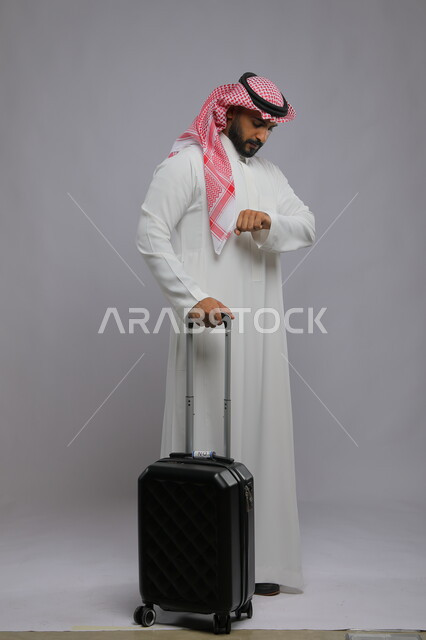 النظر إلى ساعه اليد للتاكد من الوقت ، قضاء وقت ممتع في السياحه بالعطلة الصيفيه, التأخر على موعد الطائرة، بورتريه لشاب عربي خليجي سعودي يرتدي الشماغ والثوب التقليدي يمسك بيده حقيبة سفر ،صورة الجسم بالطول الكامل ، خلفية رمادية
