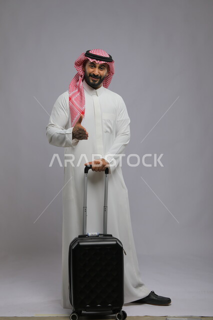النظر إلى الكاميرا بإيماءات الترحيب والمصافحة و السرور، الاستمتاع في السياحه بأوقات الفراغ، بورتريه لشاب عربي خليجي سعودي مبتسم يرتدي الشماغ والثوب التقليدي يمسك بيده حقيبة سفر ،صورة الجسم بالطول الكامل ، خلفية رمادية