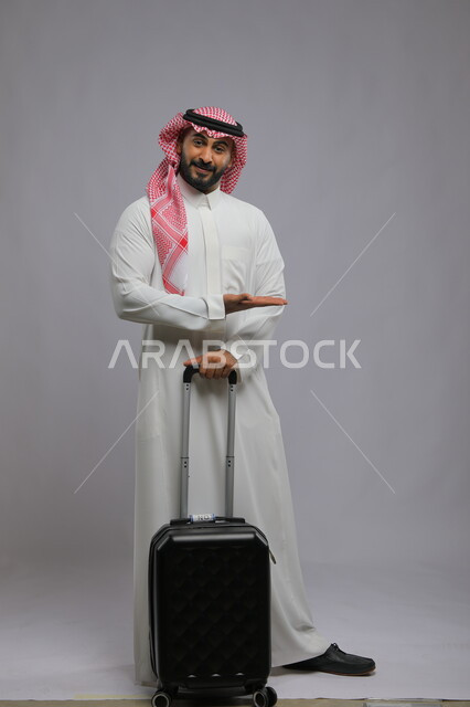 Looking at the camera with happy gestures, enjoying leisure time tourism, portrait of a smiling Saudi Gulf Arab young man wearing a shemagh and traditional thobe holding a suitcase and pointing his hand to point somewhere, full length body portrait, gray backgroundLooking at the camera with happy gestures, enjoying leisure time tourism, portrait of a smiling Saudi Gulf Arab young man wearing a shemagh and traditional thobe holding a suitcase and pointing his hand to point somewhere, full length 