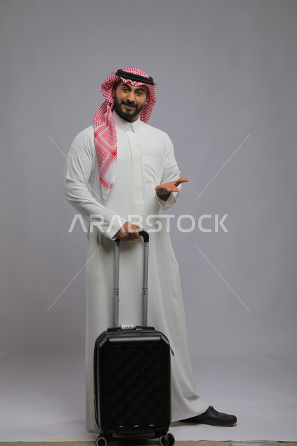 استلام جواز السفر من موظف الريسبشين ،الوقوف باستقامة والنظر إلى الكاميرا بإيماءات السرور ،قضاء وقت ممتع في السياحه بالعطلة الصيفيه، بورتريه لشاب عربي خليجي سعودي مبتسم يرتدي الشماغ والثوب التقليدي يمسك بيده حقيبة سفر ،صورة الجسم بالطول الكامل ، خلفية رمادية