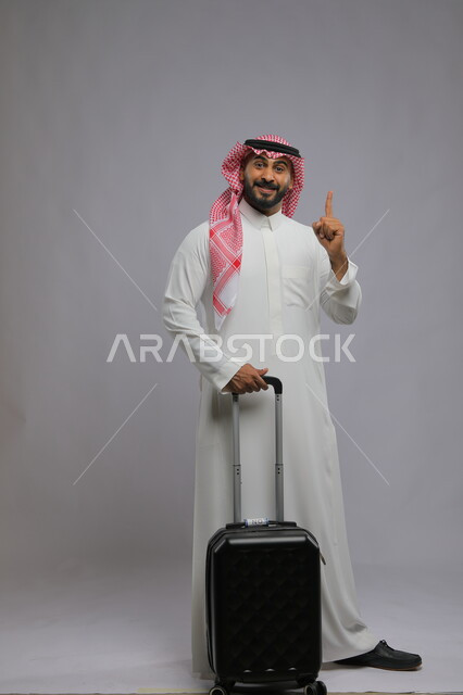 الاشارة بأصبع السبابه نحو الأعلى بإيماءات السرور ، النظر إلى الكاميرا، الاستمتاع في السياحه بأوقات الفراغ، بورتريه لشاب عربي خليجي سعودي مبتسم يرتدي الشماغ والثوب التقليدي يمسك بيده حقيبة سفر ،صورة الجسم بالطول الكامل ، خلفية رمادية