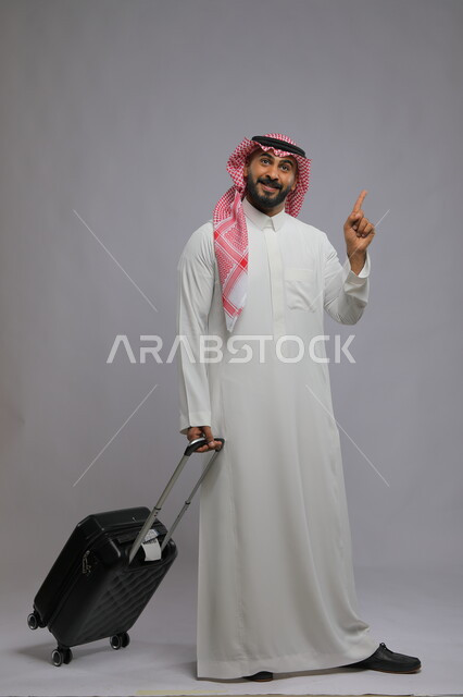  تلبية النداء والتوجه نحو الطائرة وبدء الرحلة ،الاستمتاع في السياحه بأوقات الفراغ، بورتريه لشاب عربي خليجي سعودي مبتسم يرتدي الشماغ والثوب التقليدي يمسك بيده حقيبة سفر ويشير بأصبعه لشيء ما ،صورة الجسم بالطول الكامل ، خلفية رمادية