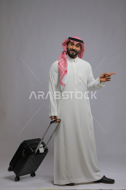 الاشارة لشيء ما بأصبع السبابة، الذهاب والمشي إلى المطار، النظر إلى الكاميرا بإيماءات السرور ،قضاء وقت ممتع في السياحه بالعطلة الصيفيه، بورتريه لشاب عربي خليجي سعودي يرتدي الشماغ والثوب التقليدي يمسك بيده حقيبة السفر ،صورة الجسم بالطول الكامل ، خلفية رمادية