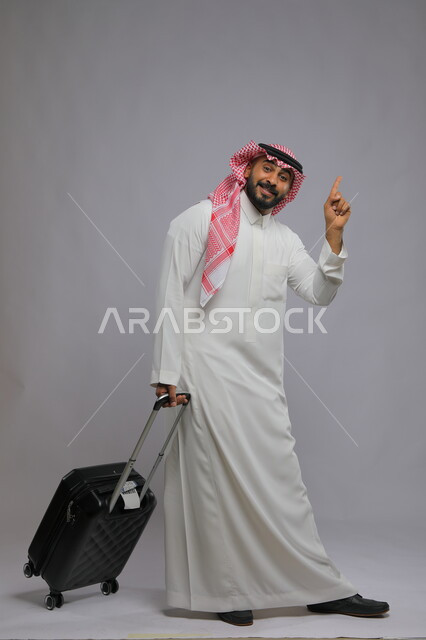 الاستمتاع في السياحه بأوقات الفراغ، رفع الاصبع بإيماءات سماع النداء للتوجه نحو الطائرة وبدء الرحلة ،بورتريه لشاب عربي خليجي سعودي مبتسم يرتدي الشماغ والثوب التقليدي يمسك بيده حقيبة سفر ،صورة الجسم بالطول الكامل ، خلفية رمادية
