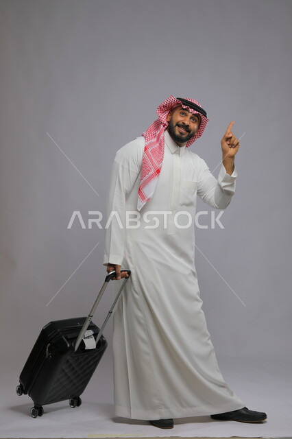 رفع الاصبع بإيماءات سماع النداء للتوجه نحو الطائرة وبدء الرحلة ،الاستمتاع في السياحه بأوقات الفراغ، بورتريه لشاب عربي خليجي سعودي مبتسم يرتدي الشماغ والثوب التقليدي يمسك بيده حقيبة سفر ،صورة الجسم بالطول الكامل ، خلفية رمادية