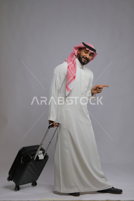 الذهاب والمشي إلى المطار، النظر إلى الكاميرا بإيماءات السرور ،قضاء وقت ممتع في السياحه بالعطلة الصيفيه، بورتريه لشاب عربي خليجي سعودي يرتدي الشماغ والثوب التقليدي يمسك بيده حقيبة السفر ويشير بأصبع السبابة لشيء ما ،صورة الجسم بالطول الكامل ، خلفية رمادية