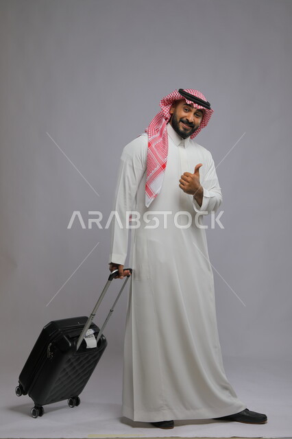 الاستمتاع في السياحه بأوقات الفراغ، إيماءات تدل على علامة الجودة والقبول والرضا ، بورتريه لشاب عربي خليجي سعودي مبتسم يرتدي الشماغ والثوب التقليدي يمسك بيده حقيبة سفر وينظر إلى الكاميرا ،صورة الجسم بالطول الكامل ، خلفية رمادية