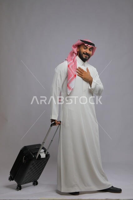 النظر إلى الكاميرا بتعابير الشكر والترحيب ،الاستمتاع في السياحه بأوقات الفراغ، بورتريه لشاب عربي خليجي سعودي مبتسم يرتدي الشماغ والثوب التقليدي يمسك بيده حقيبة سفر ،صورة الجسم بالطول الكامل ، خلفية رمادية
