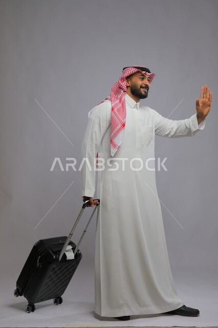ايماءات الاستقبال عند الوصول، الوداع والانطلاق لبدء الرحلة، قضاء وقت ممتع في السياحه بالعطلة الصيفيه ،بورتريه لشاب عربي خليجي سعودي يرتدي الشماغ والثوب التقليدي يمسك حقيبة السفر ،صورة الجسم بالطول الكامل ، خلفية رمادية