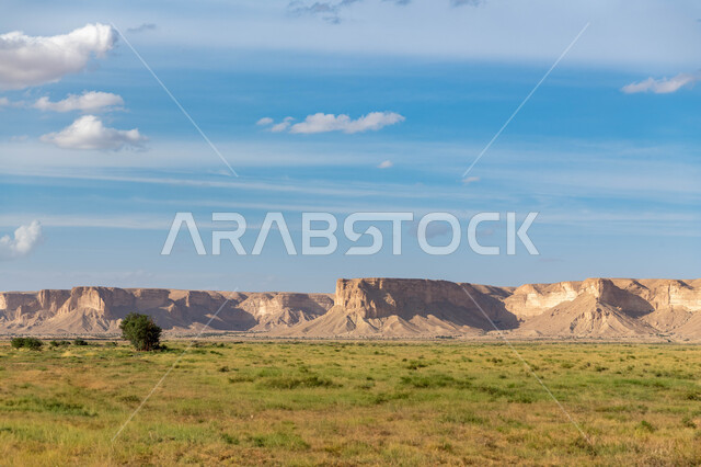 سلسلة جبال طويق في الرياض في المملكه العربيه السعوديه, مطل الجراند كانيون حافة نهاية العالم, مناطق سياحيه مميزه في الرياض, منظر الغيوم في السماء الزرقاء، النباتات ومساحات المروج الخضراء.