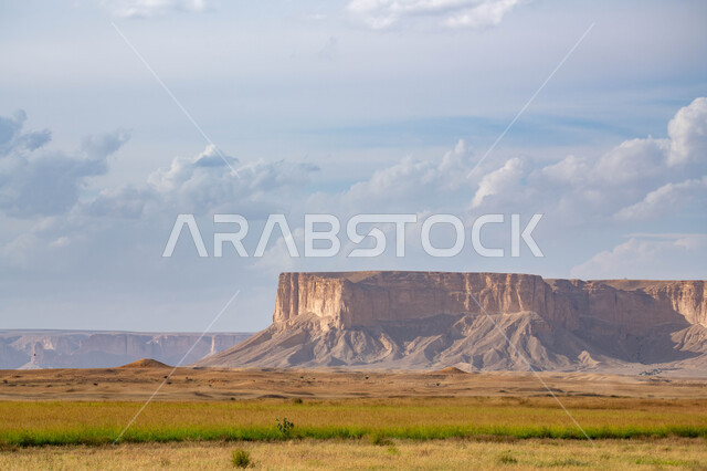 سلسلة جبال طويق في الرياض في المملكه العربيه السعوديه, مطل الجراند كانيون حافة نهاية العالم, مناطق سياحيه مميزه في الرياض, منظر الغيوم في السماء الزرقاء، النباتات ومساحات المروج الخضراء.