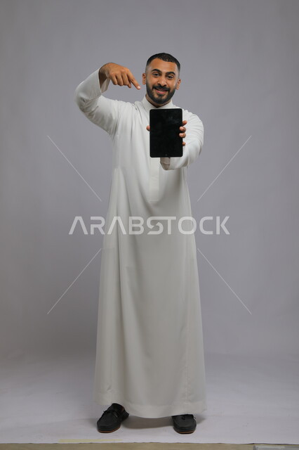 التسويق الإلكتروني وعرض خدمات وتطبيقات الجوال، بورتريه لرجل عربي سعودي خليجي يرتدي الزي التقليدي ويشير بيده إلى جهاز التابلت بشاشه سوداء فارغه، ايماءت الاعجاب باستخدام التقنيه والتكنولوجيا الحديثه، صورة الجسم بالطول الكامل، خلفية رمادية