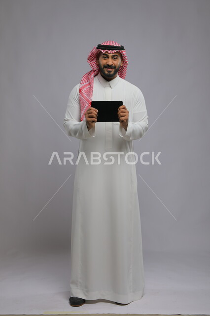 Displaying a blank black screen on the tablet, using modern electronic digital marketing technologies, liking tablet applications and services, a portrait of a smiling Gulf Arab Saudi man wearing a shemagh and traditional thobe looking at the camera, full-length body photo, gray background