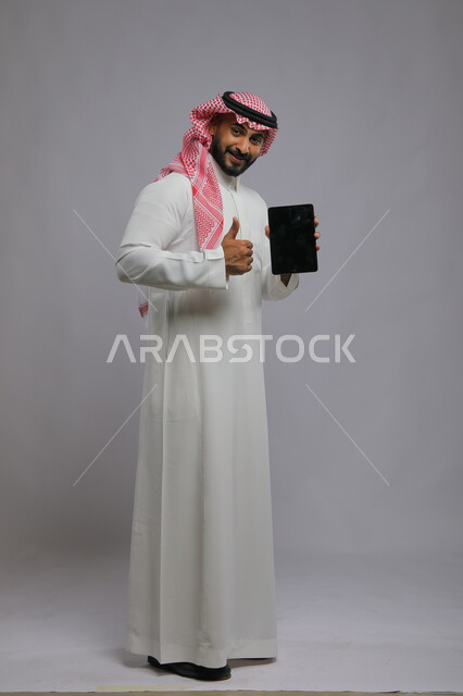 Displaying a blank black screen on the tablet, using modern electronic digital marketing technologies, the quality of tablet applications and services, a portrait of a smiling Saudi Arabian Gulf man wearing a shemagh and traditional thobe, looking at the camera with admiring gestures, full-length body photo, gray background