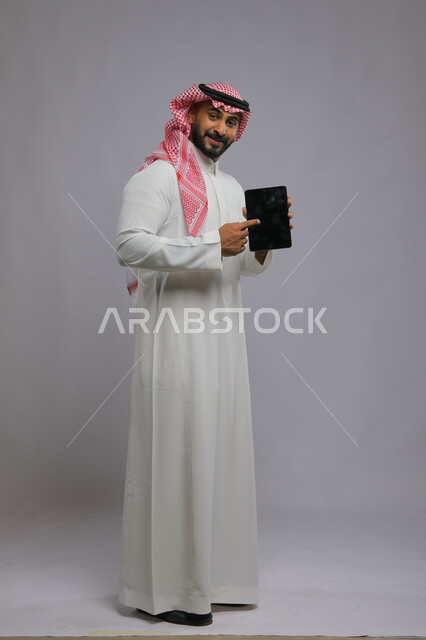 الاشارة إلى التابلت بشاشة سوداء فارغة، استعمال التقنيات الحديثة للتسويق الرقمي الالكتروني، عرض تطبيقات وخدمات الجهاز اللوحي ، بورتريه لشاب عربي خليجي سعودي مبتسم يرتدي الشماغ و الثوب التقليدي ينظر إلى الكاميرا ،صورة الجسم بالطول الكامل، خلفية رمادية