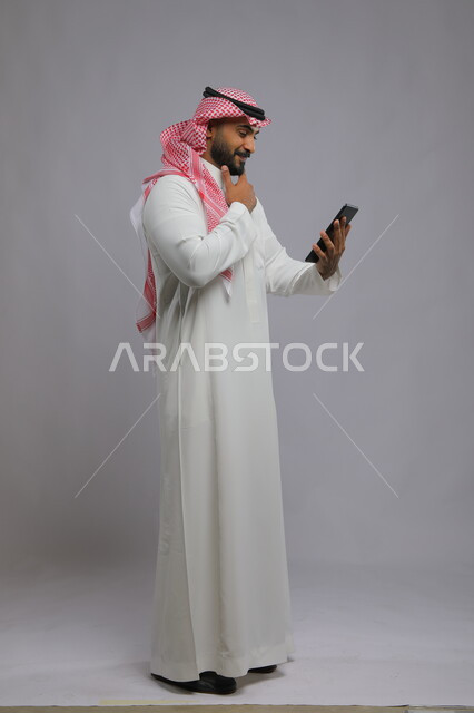 الدردشة مع الأهل والأقارب، مشاهدة البرامج المفضلة بإيماءات التفكير ,استخدام التكنولوجيا و التقنيات الحديثة، بورتريه لرجل عربي خليجي سعودي يرتدي الشماغ و الثوب التقليدي يقوم بإجراء مكالمة فيديو عبر جهاز التابليت ،صورة الجسم بالطول الكامل, خلفية رمادية