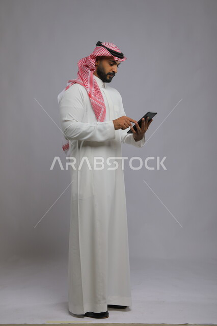تصفح برامج التواصل الاجتماعي ،متابعة آخر أخبار السوشال ميديا ،استخدام التكنولوجيا و التقنيات الحديثة، بورتريه لرجل عربي خليجي سعودي يرتدي الشماغ و الثوب التقليدي يحمل بيده جهاز التابليت، تطبيقات وبرامج الأجهزة اللوحية، صورة الجسم بالطول الكامل ,خلفية رمادية