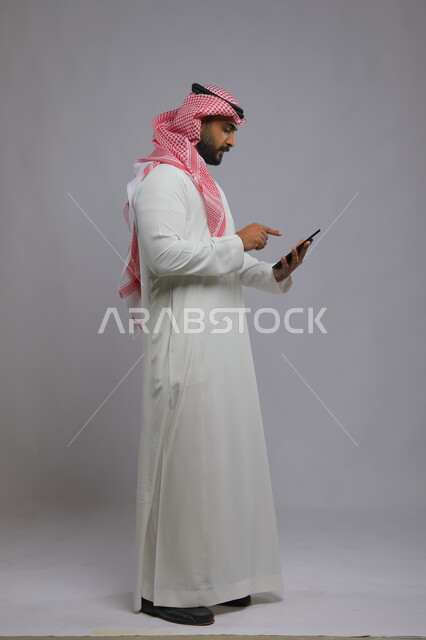 Following the latest social media news, using technology and modern techniques, a portrait of a Saudi Gulf Arab man wearing a shemagh and traditional dress, holding a tablet in his hand, browsing social media programs, tablet applications and programs, full-length body photo, gray background