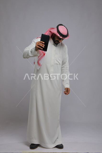 ايماءات الحزن والخساره بعد مشاهدة شيء ما على التابليت ،استخدام التكنولوجيا و التقنيات الحديثة، بورتريه لرجل عربي خليجي سعودي يرتدي الشماغ و الثوب التقليدي يشعر بالانزعاج والغضب وينظر الى الأسفل ، صورة الجسم بالطول الكامل, خلفية رمادية