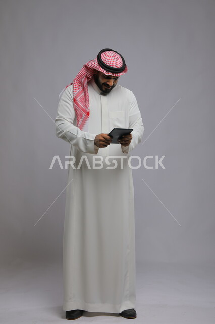 صورة الجسم بالطول الكامل, مشاهدة البرامج المفضلة بإيماءات الغضب والاستياء ,استخدام التكنولوجيا و التقنيات الحديثة، بورتريه لرجل عربي خليجي سعودي يرتدي الشماغ و الثوب التقليدي يحمل بيده جهاز التابليت و يقوم بإجراء مكالمة فيديو ،خلفية رمادية