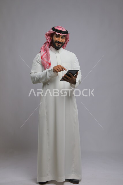 Chatting with family and friends, looking at the camera with happy gestures, using technology and modern techniques, portrait of a smiling Saudi Arabian Gulf man wearing a shemagh and traditional thobe, pointing with his finger on the tablet, full-length body portrait, gray background