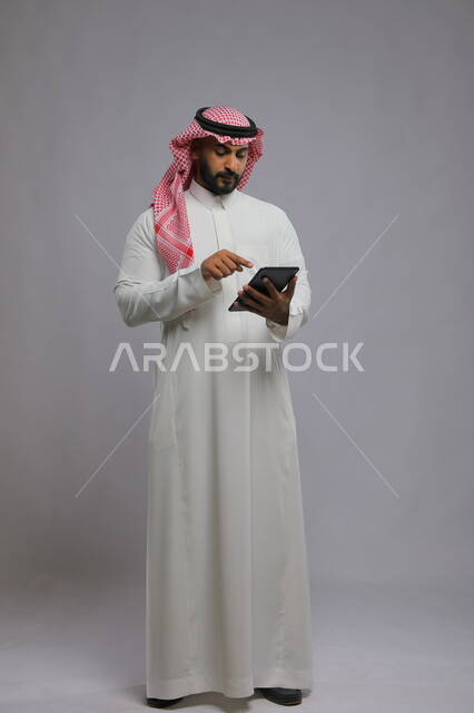 Browsing social networking sites, using technology and modern technologies, a portrait of a Saudi Gulf Arab man wearing a shemagh and traditional thobe, tapping his finger on a tablet device, chatting with family and friends, tablet applications and programs, full-length body image, gray background