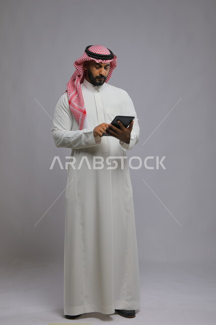 استخدام التكنولوجيا و التقنيات الحديثة , بورتريه لرجل عربي خليجي سعودي يرتدي الشماغ و الثوب التقليدي يحمل بيده جهاز التابليت ،الدردشة مع الأهل والأصدقاء , تصفح برامج التواصل الاجتماعي ، تطبيقات وبرامج الأجهزة اللوحية، صورة الجسم بالطول الكامل , خلفية رمادية .