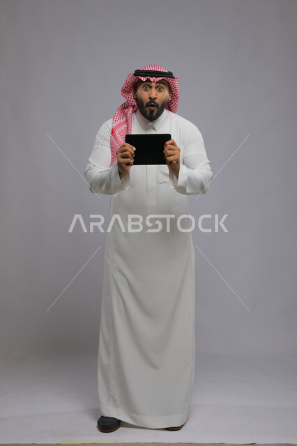 Gestures of emotion and engagement while watching a football match, displaying a blank black advertising screen on the tablet, enthusiasm and astonishment at victory and triumph, portrait of a Saudi Gulf Arab young man wearing a shemagh and traditional thobe looking at the camera, full-length body photo, gray background to see