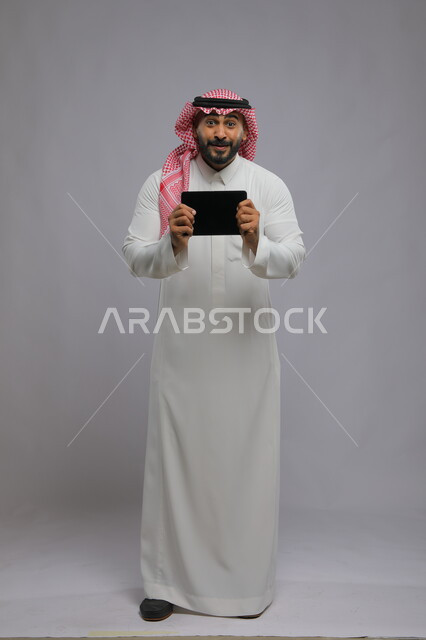 عرض شاشة اعلانية سوداء فارغه على الجهاز اللوحي، ايماءات الانفعال والاندماج أثناء مشاهدة مباراة كرة القدم،  الحماس والدهشه بالفوز والنصر ، بورتريه لشاب عربي خليجي سعودي يرتدي الشماغ و الثوب التقليدي ينظر إلى الكاميرا ،صورة الجسم بالطول الكامل، خلفية رمادية