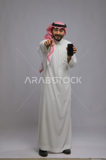 استخدام التقنيات الحديثة لإنجاز المهام اليومية ،الاشارة بالاصبع إلى الامام ،عرض شاشة سوداء فارغة عبر الجوال، تطبيقات وخدمات الهاتف المحمول، بورتريه لشاب عربي خليجي سعودي مبتسم يرتدي الشماغ و الثوب التقليدي ينظر إلى الكاميرا ،صورة الجسم بالطول الكامل، خلفية رمادية