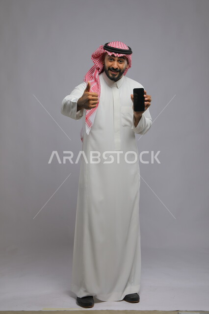 ايماءات الجودة والاعجاب بتطبيقات وخدمات الهاتف المحمول ،عرض شاشة سوداء فارغة عبر الجوال، استخدام التكنولوجيا المتطورة للتسويق الالكتروني ، بورتريه لشاب عربي خليجي سعودي مبتسم يرتدي الشماغ و الثوب التقليدي ينظر إلى الكاميرا ،صورة الجسم بالطول الكامل، خلفية رمادية